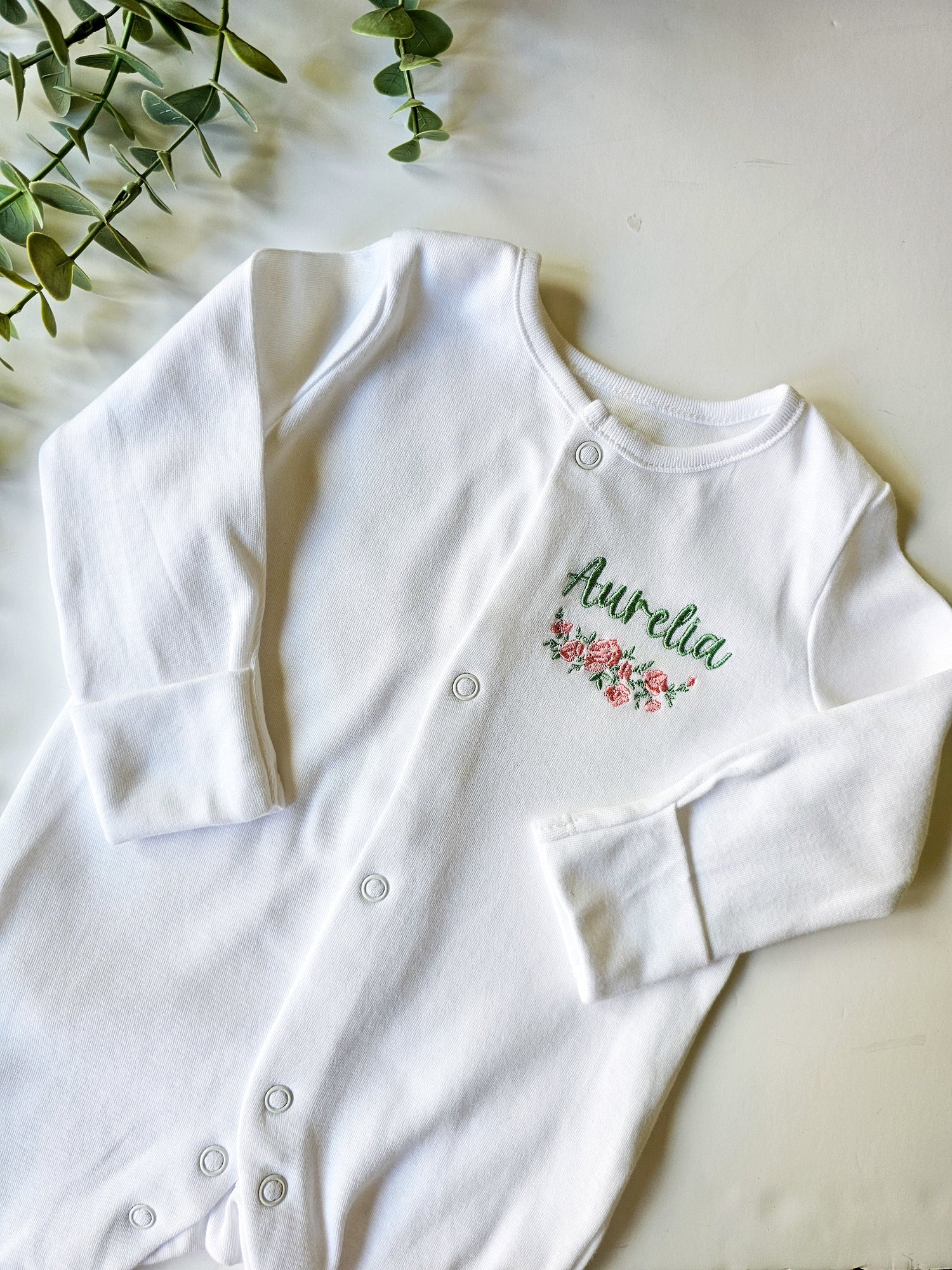 White personalized baby sleep suit, embroidered with pink and green flowers underlining baby's name in green swirly font.