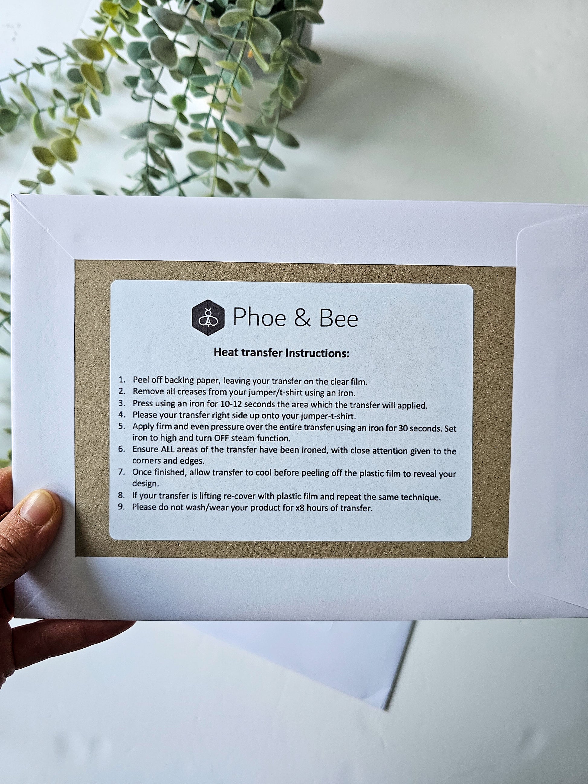 Hand holding a paper with 'Phoe & Bee' heat transfer instructions against a light background.