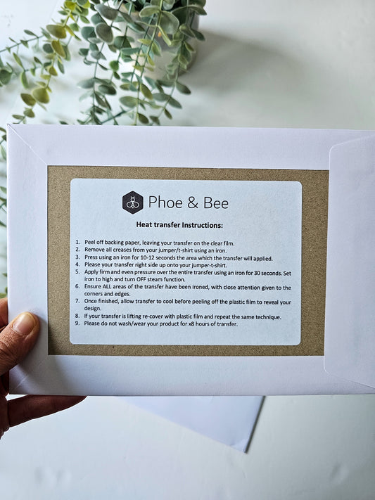 Hand holding a paper with 'Phoe & Bee' heat transfer instructions against a light background.