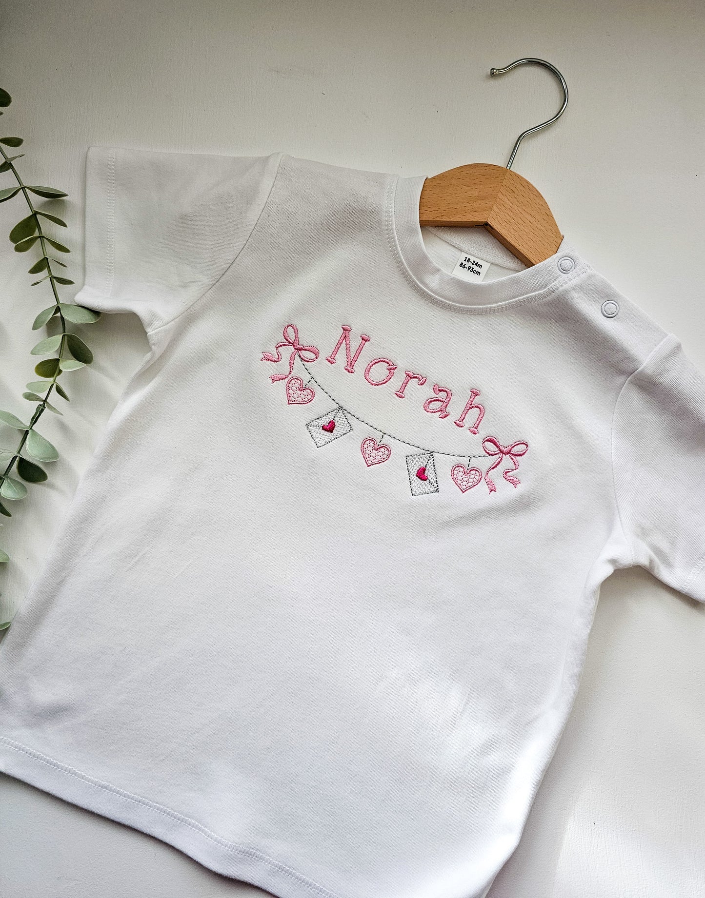 White tshirt with 'Norah' personalized embroidered and valentine bunting on a wooden hanger against a light background.