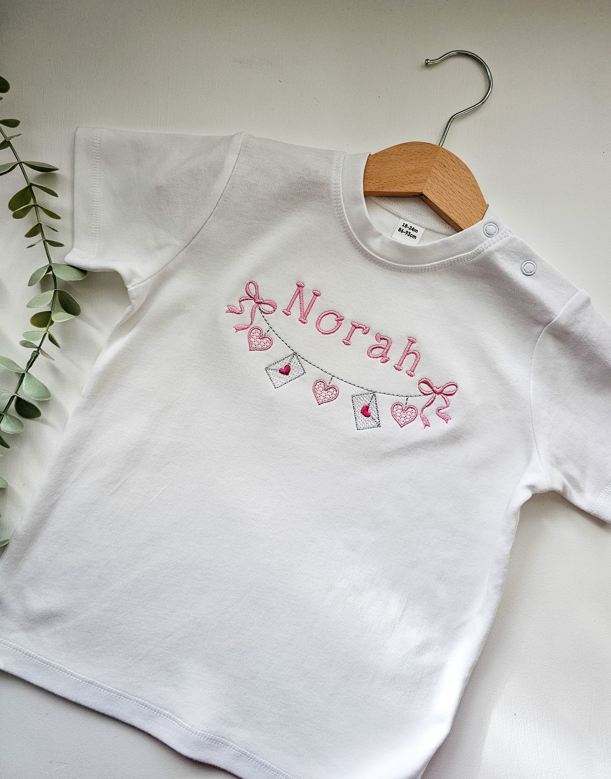 White tshirt with 'Norah' personalized embroidered and valentine bunting on a wooden hanger against a light background.