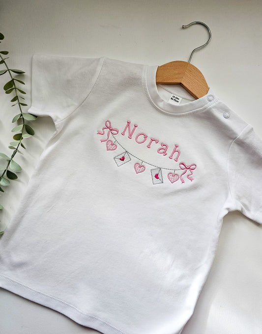 White tshirt with 'Norah' personalized embroidered and valentine bunting on a wooden hanger against a light background.