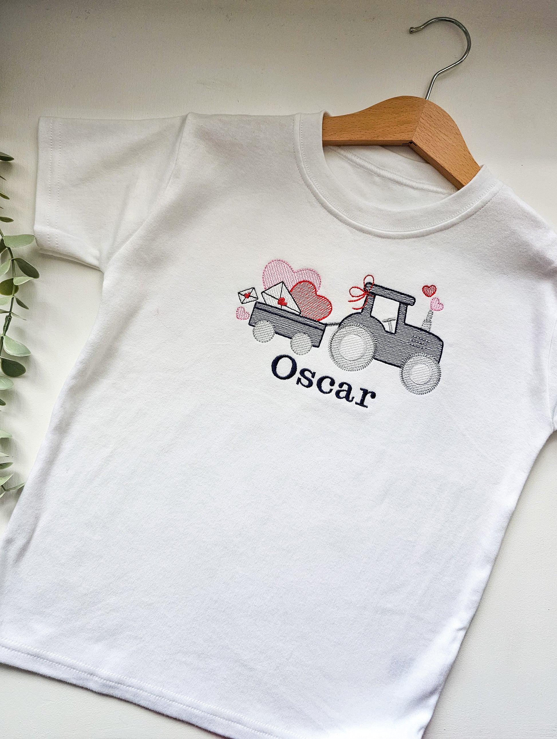 White t-shirt with a graphic of a tractor and hearts, personalized with 'Oscar', on a wooden hanger.