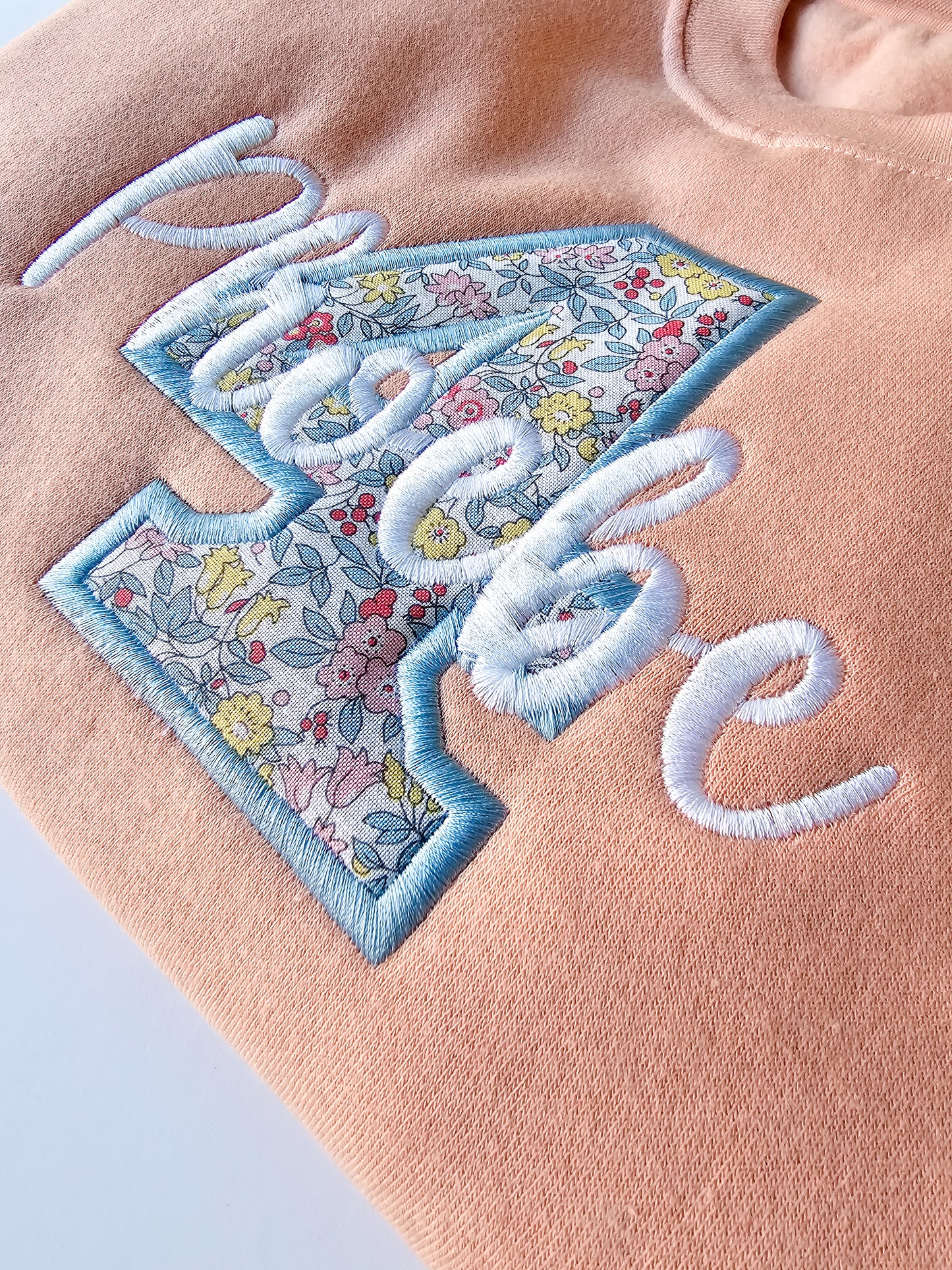 Close up of peach personalised jumper, embroidered with liberty print four and child's name in white thread.