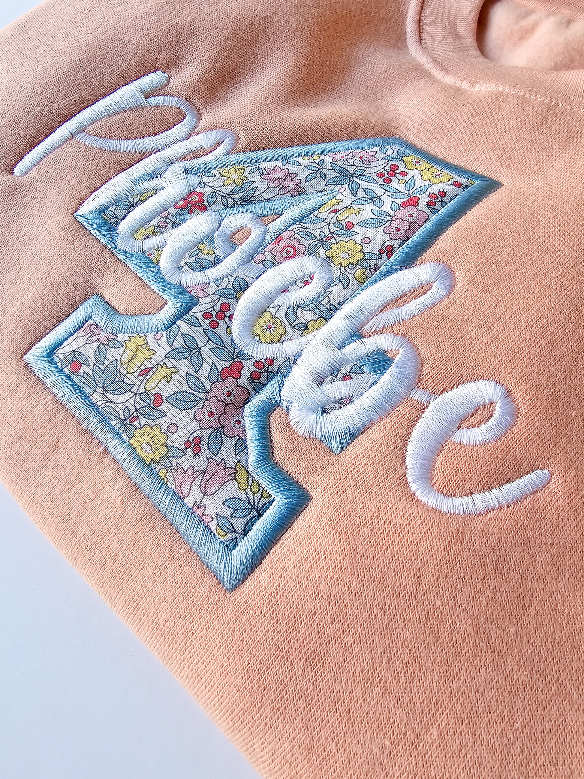 Close up of peach personalised jumper, embroidered with liberty print four and child's name in white thread.