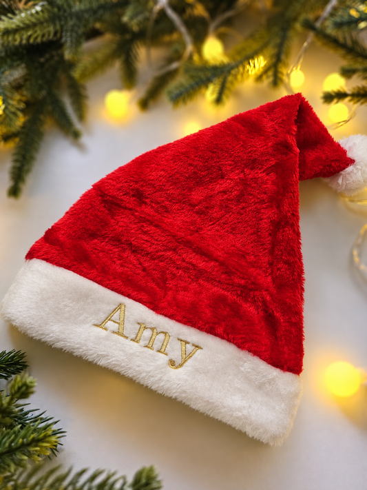 A red and white Santa hat with the name 'Amy' embroidered in gold on the white trim, styled with pine branches and warm yellow fairy lights on a white background.