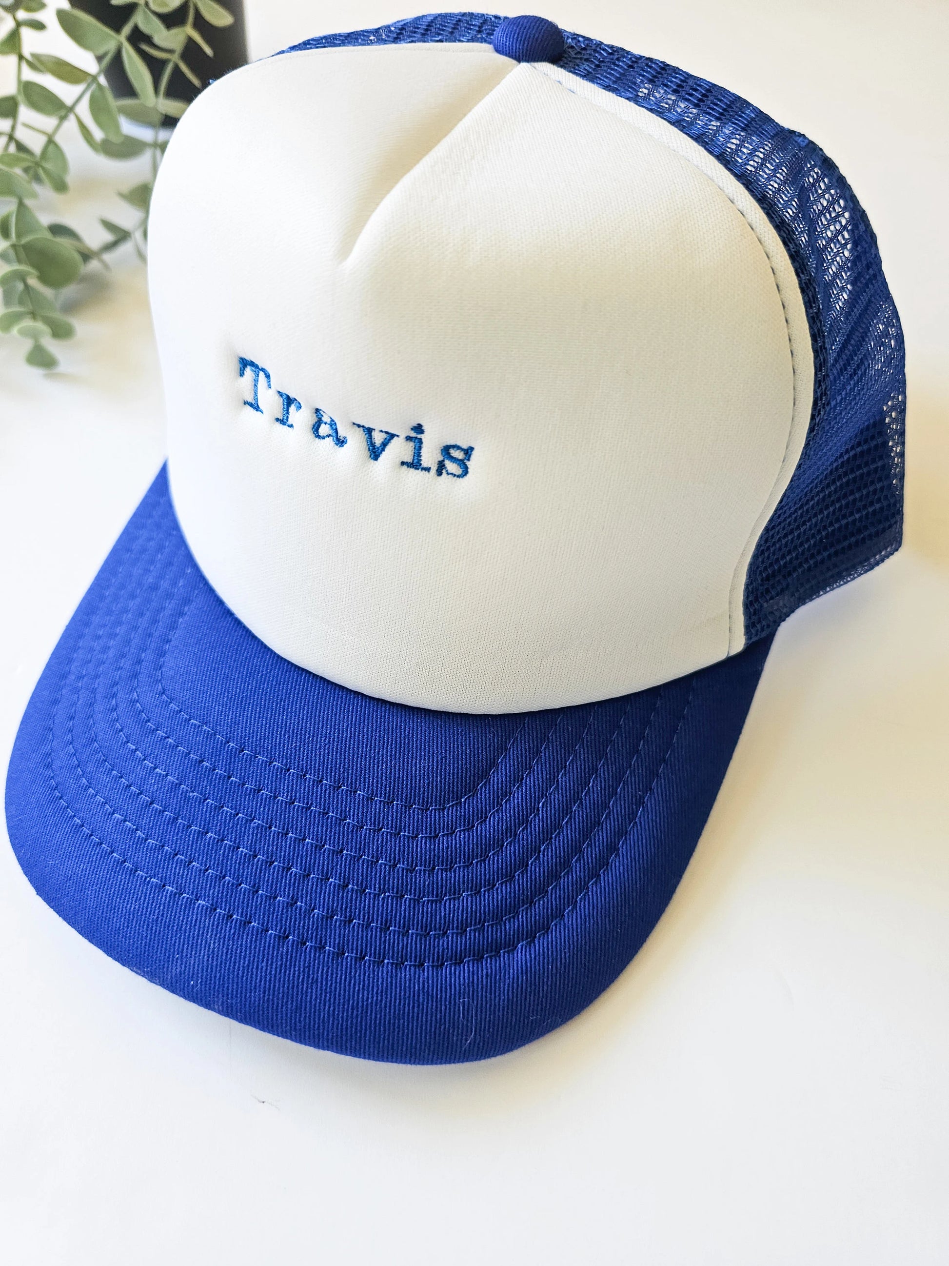 A children's trucker mesh hat in blue with white embroidery reading 'Travis' on the front.