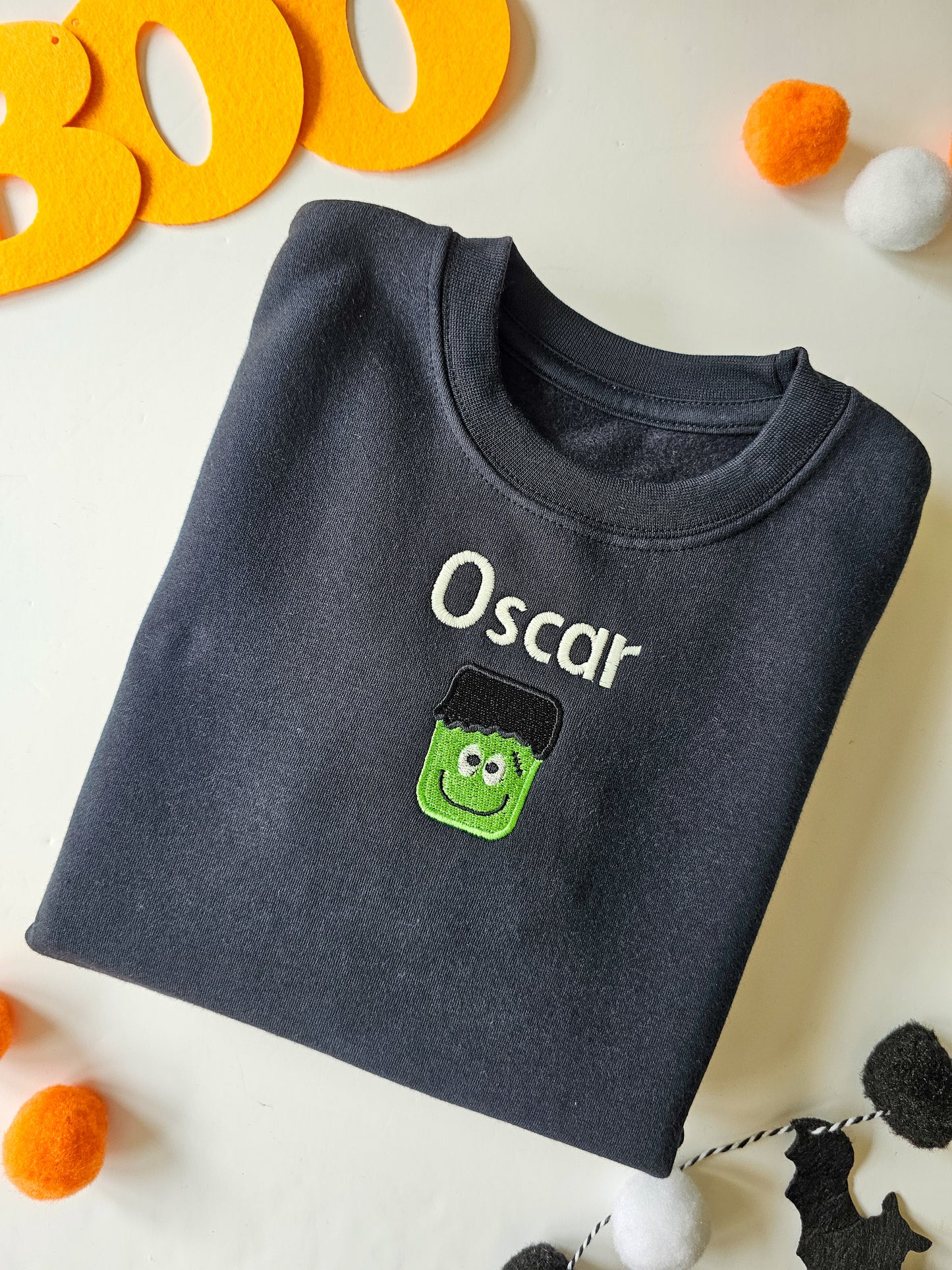 A black crew neck jumper for kids with 'OSCAR' embroidered in white. The jumper is presented on a surface with orange and black decorations and a green ghost-shaped embroidery on the front.