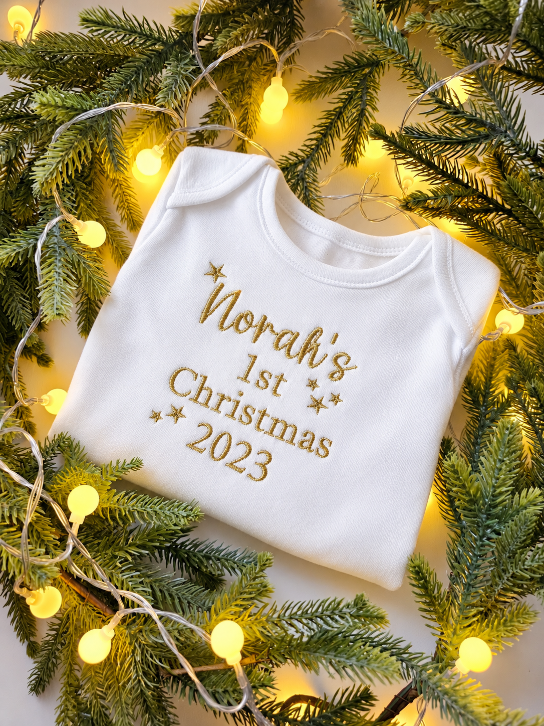 A folded white baby bodysuit with gold embroidered text reading 'Norah's 1st Christmas 2023' and small gold stars, styled on a light surface with green pine branches and warm yellow fairy lights.