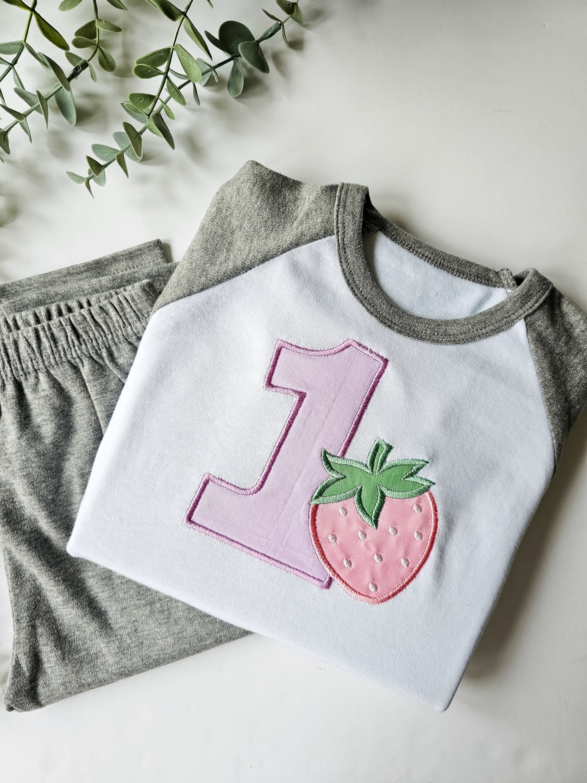 Children's birthday pyjamas with embroidered number 1 and a strawberry design on a grey background, displayed with matching pants.