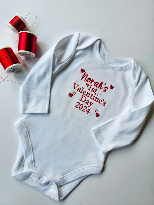 A long-sleeved white baby vest with red and gray hearts and the text 'Herah's Valentine's Day 2025' embroidered on it, accompanied by a white bib with the same design.