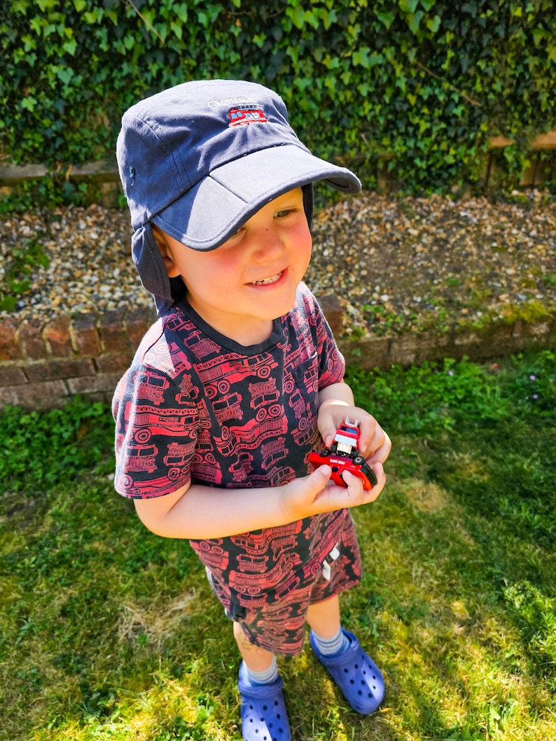 Little boy wearing a navy blue children's legionnaires cap embroidered with the name 'Oscar' and an illustration of a red fire engine on the front.