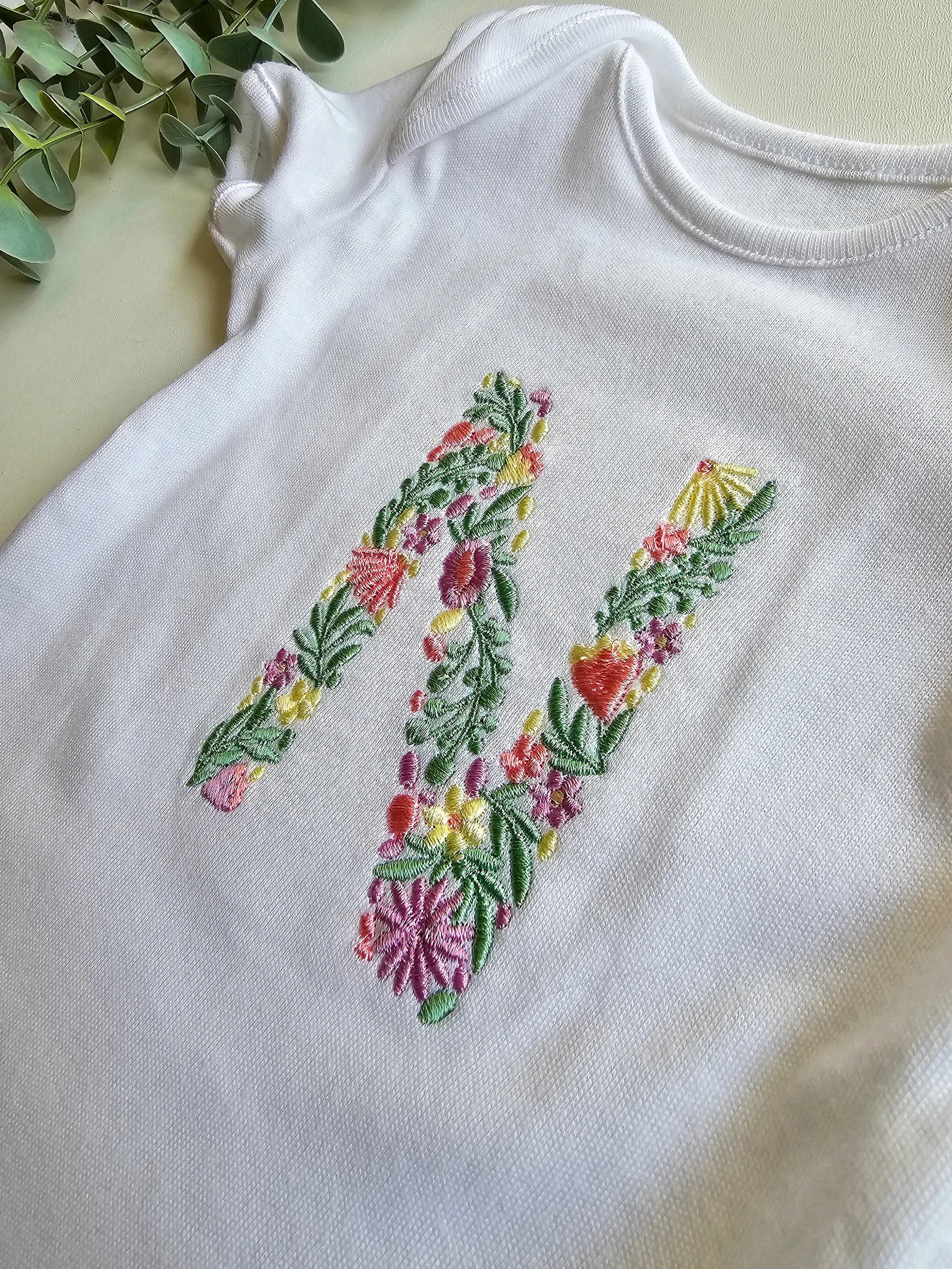 White baby onesie with floral letter 'M' embroidery on a light background