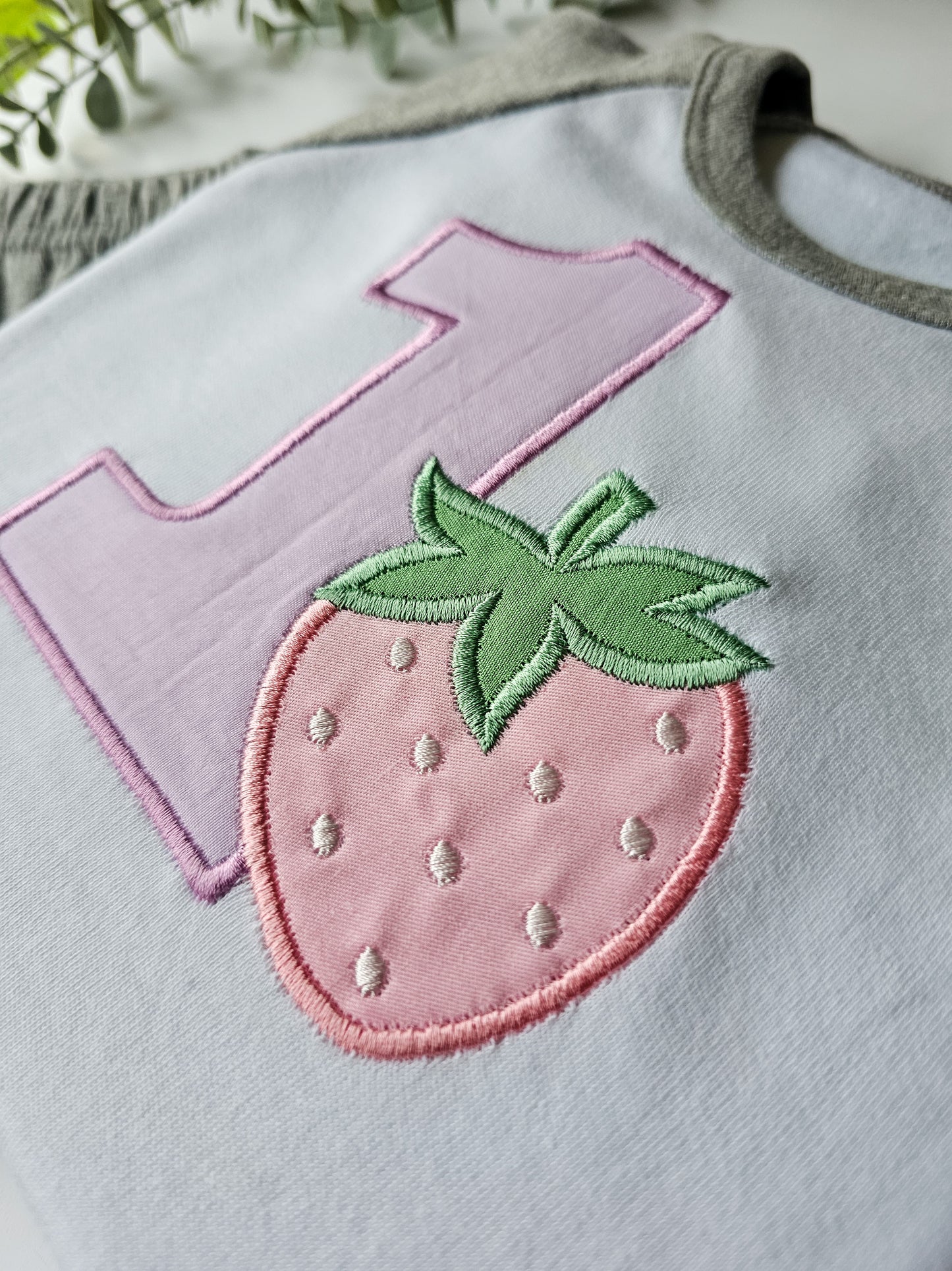close up of Children's birthday pyjamas with embroidered number 1 and a strawberry design on a grey background, displayed with matching pants.
