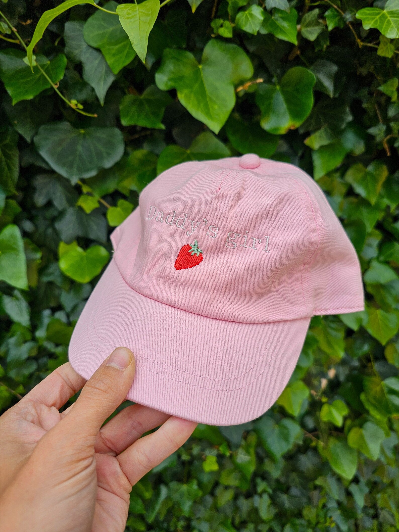 Pink cap with a strawberry embroidery personalised with daddy's girl held against a green leafy background