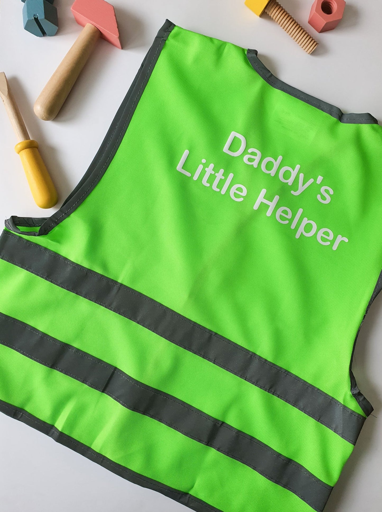 A green personalised children's high-visibility vest with 'Daddy's Little Helper' printed on the front, placed on a white surface with wooden tools and toys around it.
