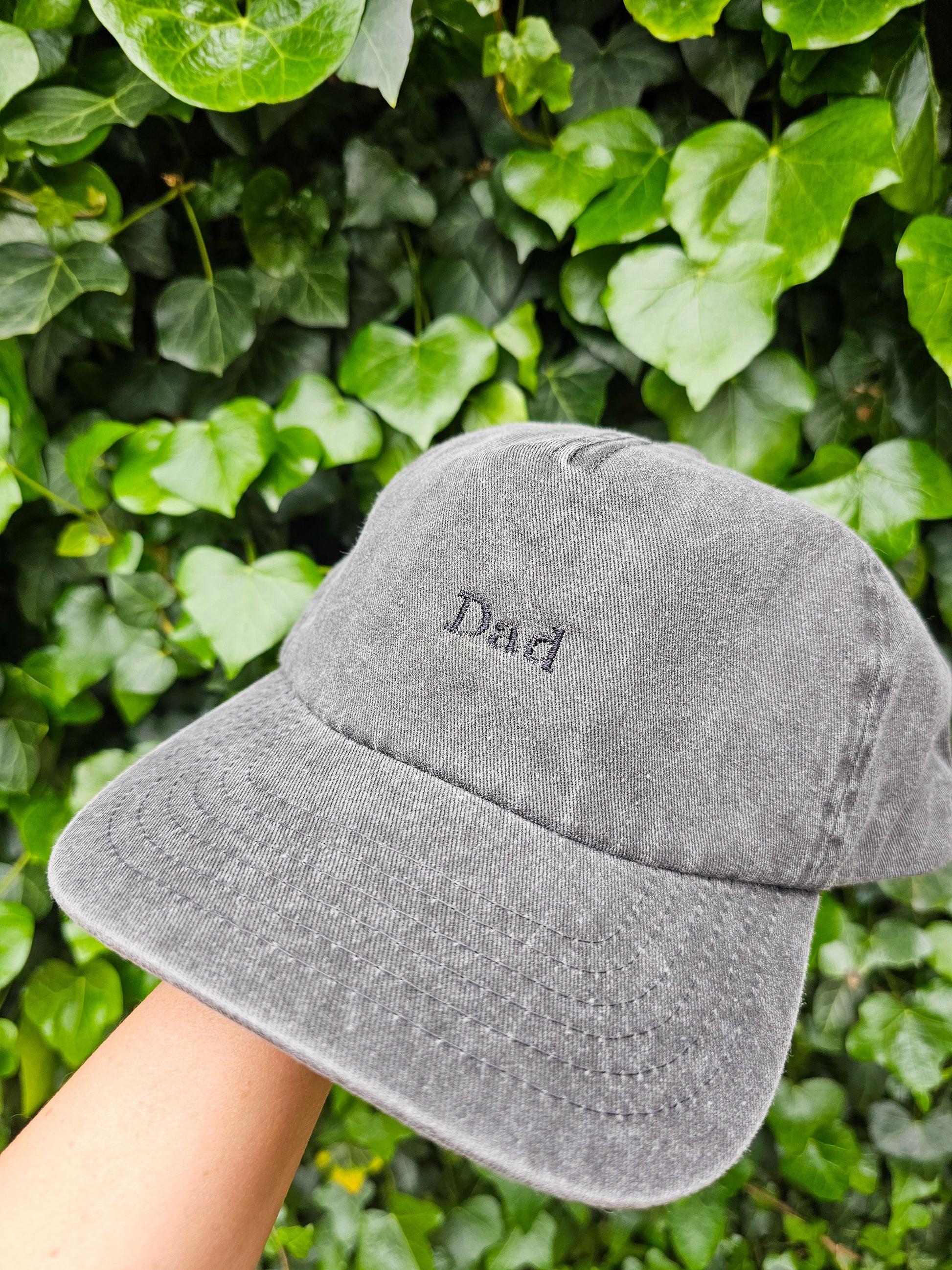 A hand holding a gray personalised embroidered cap with 'DAD' embroidered on the front, with a background of green leaves.