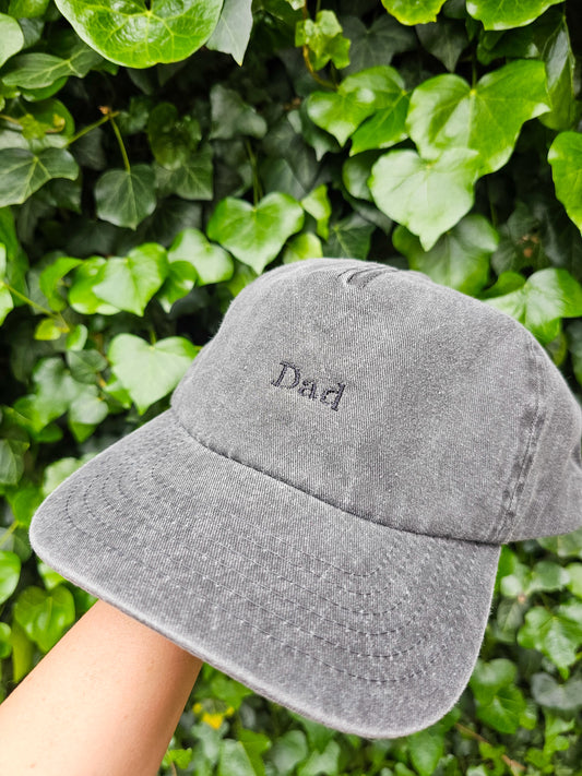 A hand holding a gray personalised embroidered cap with 'DAD' embroidered on the front, with a background of green leaves.