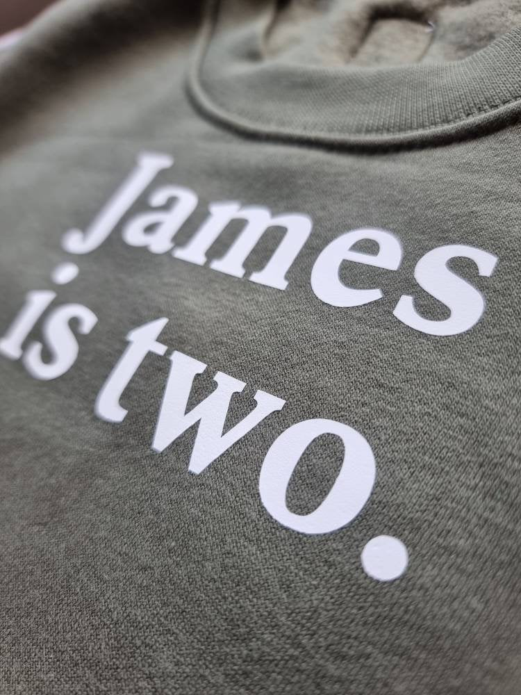 Close up of khaki kids jumper, personalised with printed james is two in white vinyl