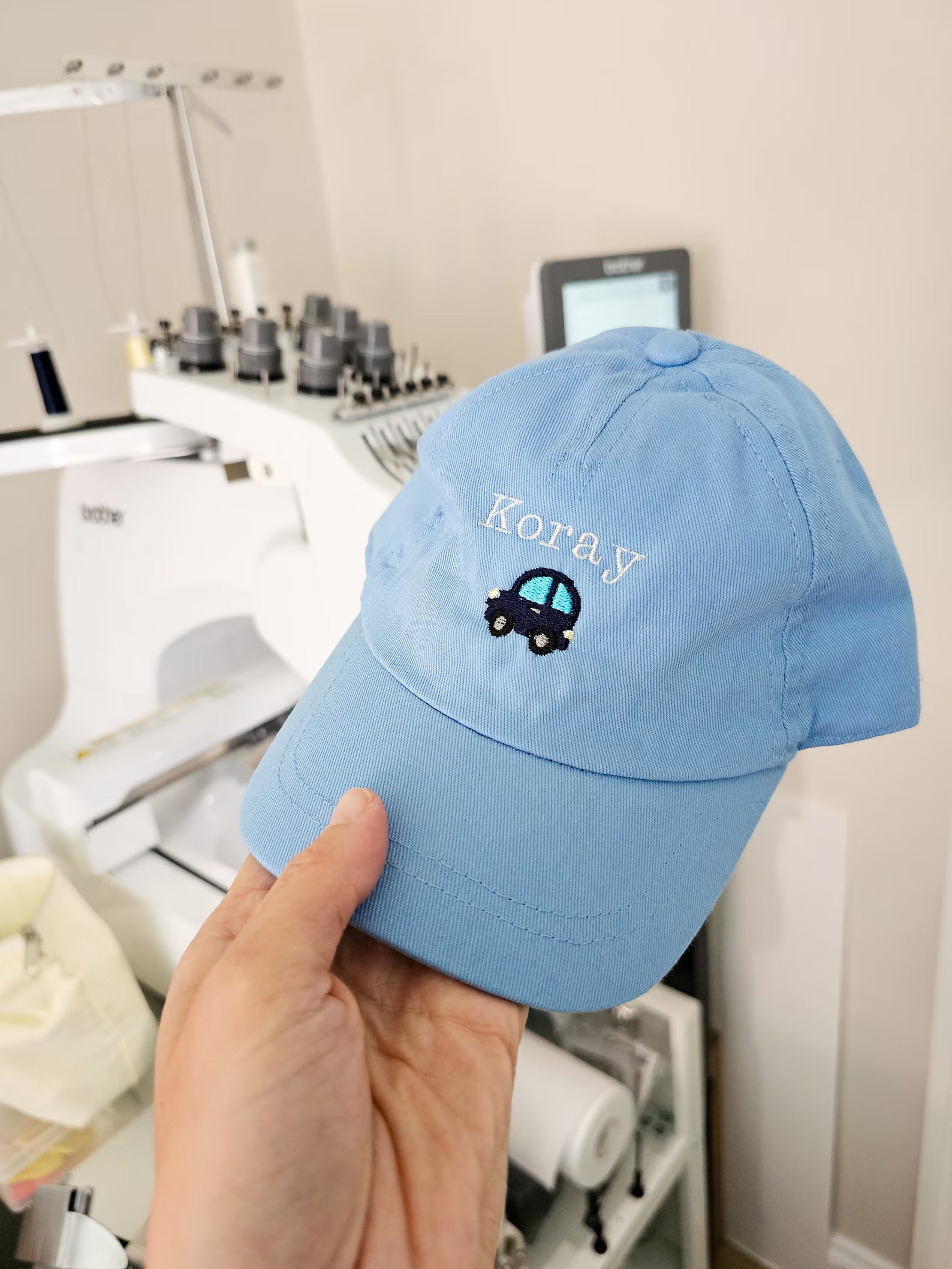 Light blue cap personalised embroidery with 'Koray' and a car logo held in front of sewing equipment.