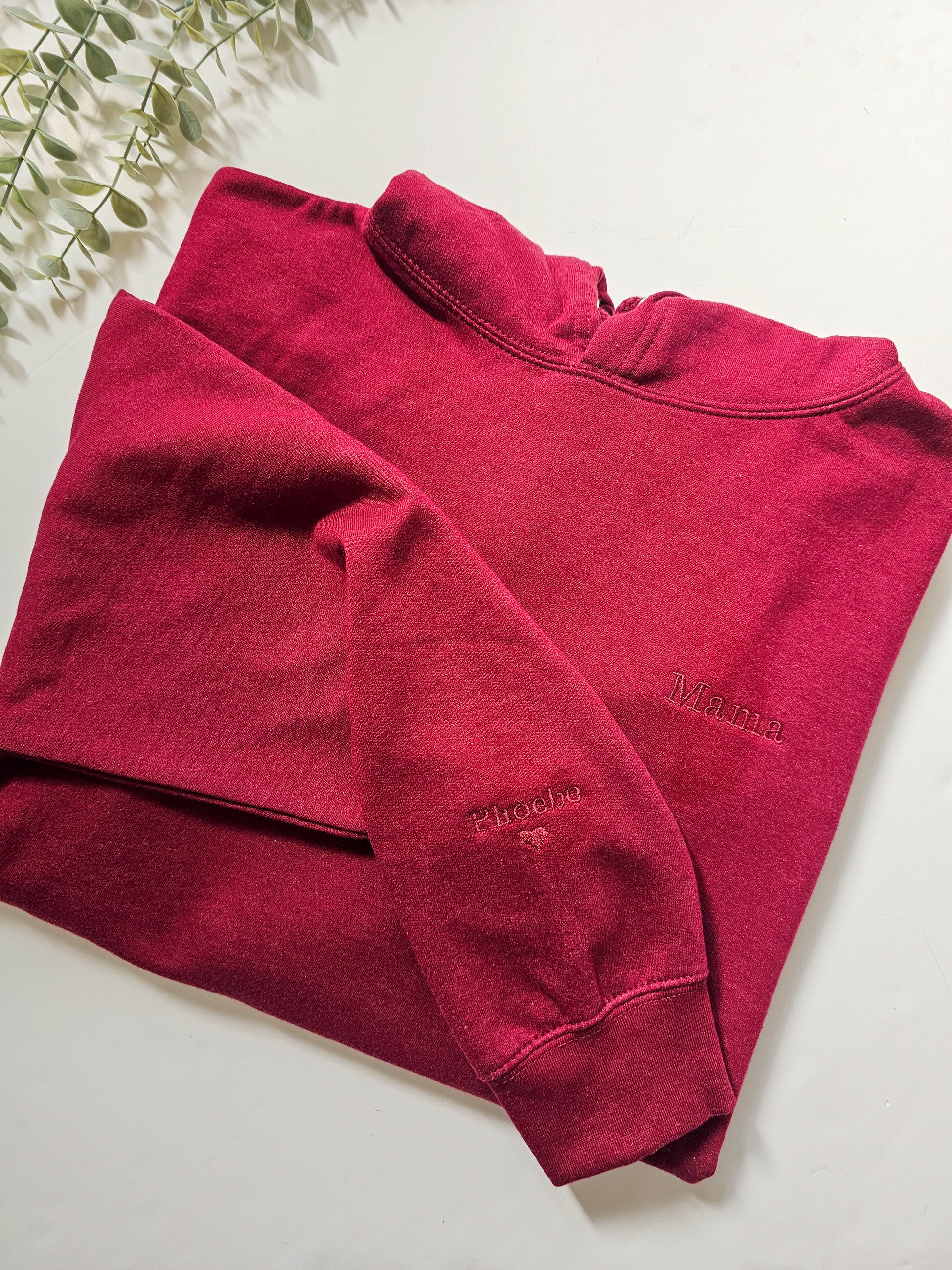 A red personalised embroidered hoodie with the name 'Mama' and a heart symbol on the sleeve, displayed on a white background.