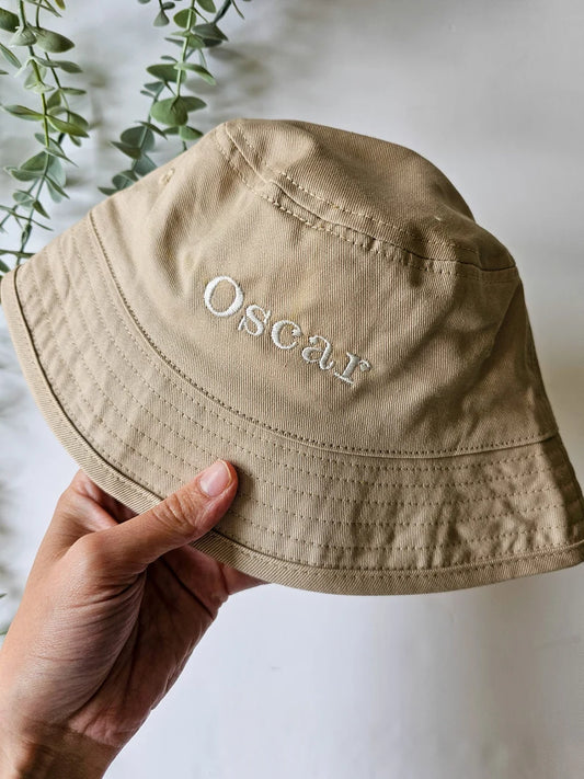 A personalised beige bucket hat with the name 'Oscar' embroidered on it, held in a hand against a background with greenery.