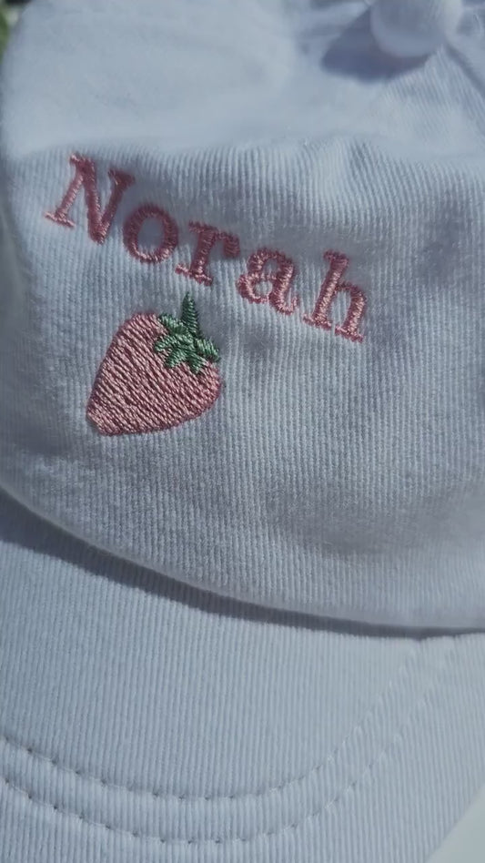 A white toddler cap with the name 'Norah' embroidered on the front, featuring a small pink strawberry below the name.