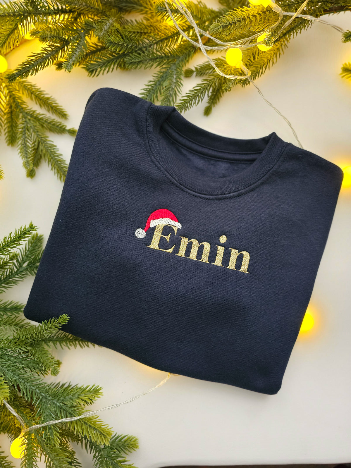 A folded navy blue sweatshirt with the name 'Emin' embroidered in gold and a small red Santa hat, styled on a white surface with pine branches and warm string lights.