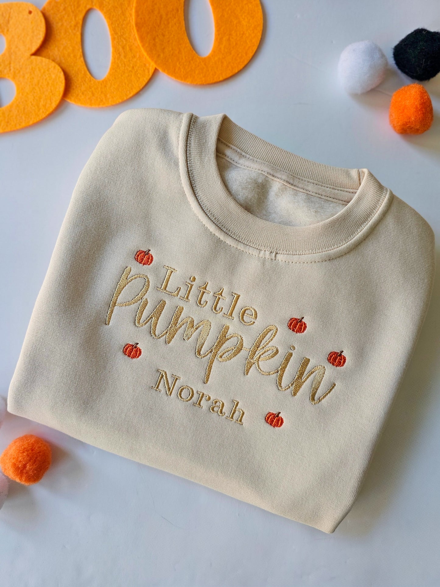 Beige baby sweater with 'Little Pumpkin Norah' text, surrounded by Halloween decorations on a white background.