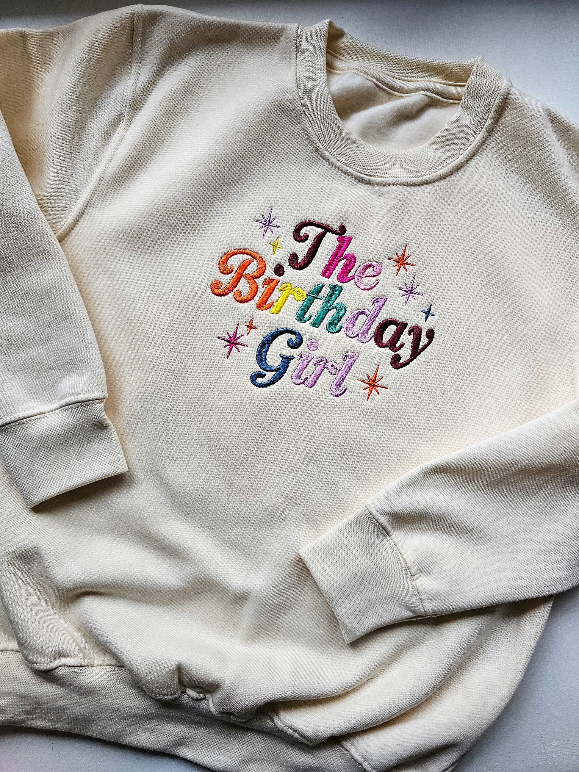 Beige sweatshirt with 'The Birthday Girl' text in colorful letters on a light gray background