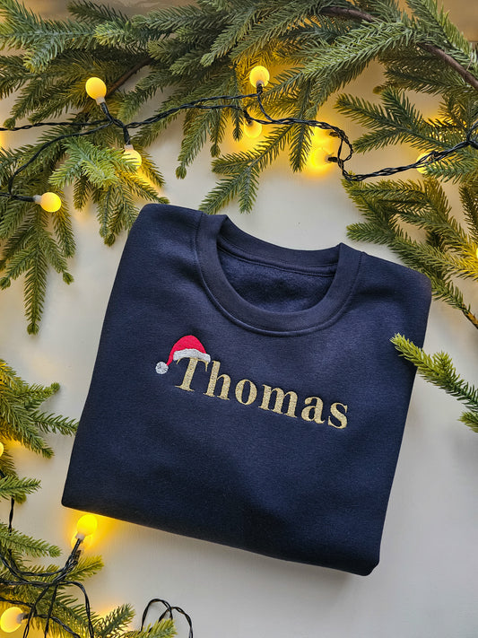 A folded navy blue crewneck sweatshirt with 'Thomas' embroidered in gold across the chest, decorated with a small red Santa hat graphic. The sweatshirt is styled in a flat lay with green pine branches and warm yellow string lights.
