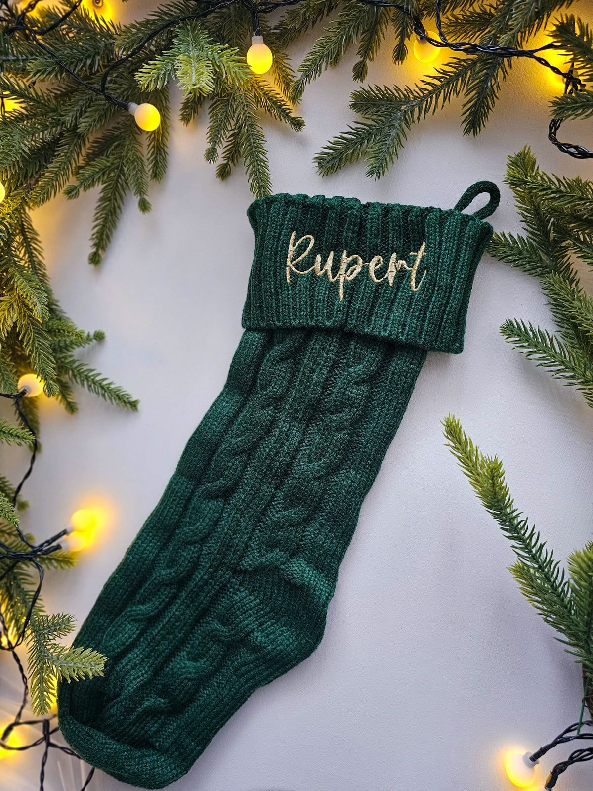 A dark green cable-knit Christmas stocking with the name 'Rupert' embroidered in white on the cuff, displayed on a white background with pine branches and warm yellow fairy lights.