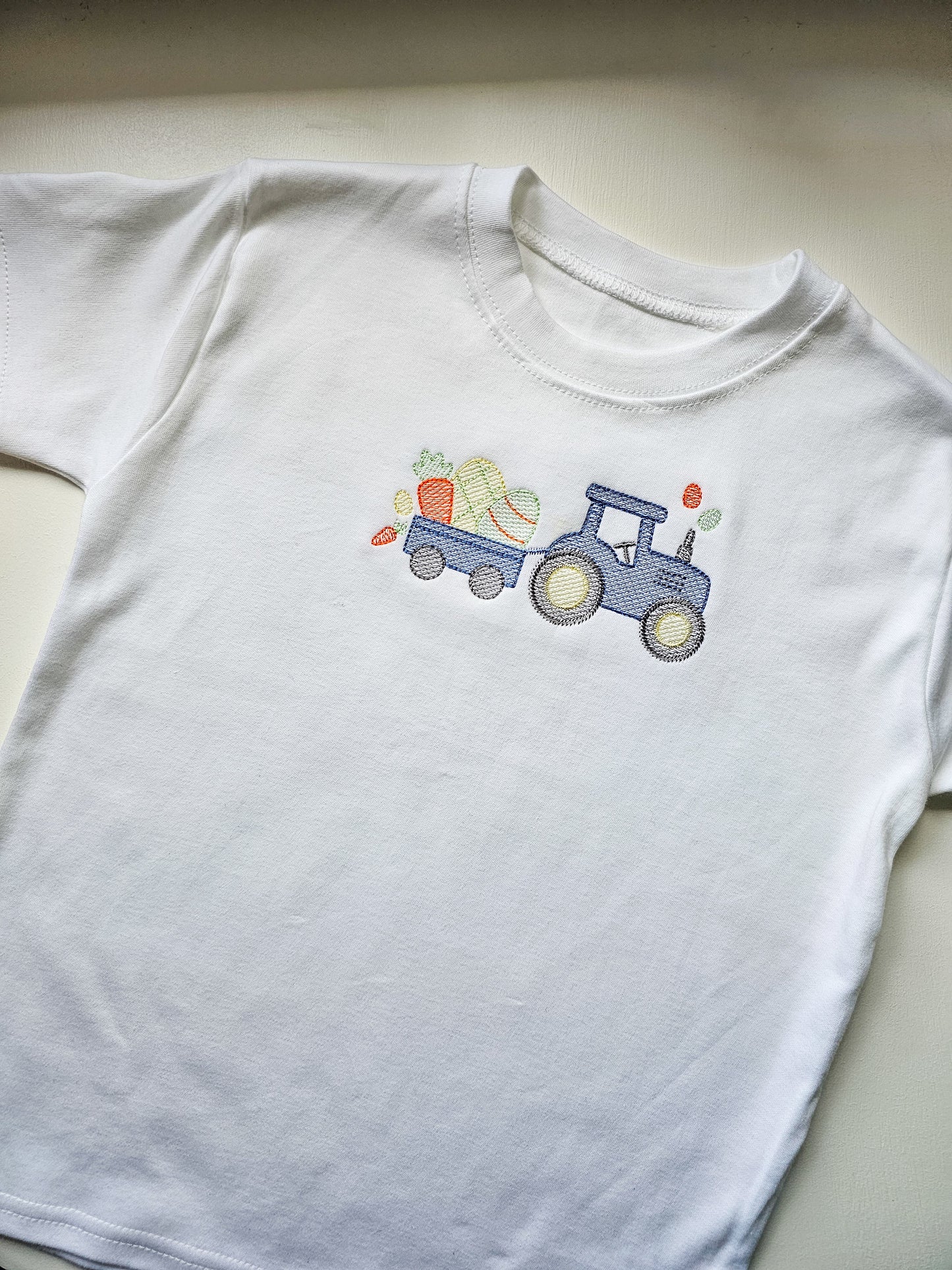 White t-shirt with a blue tractor and colorful vegetables design on a light gray background