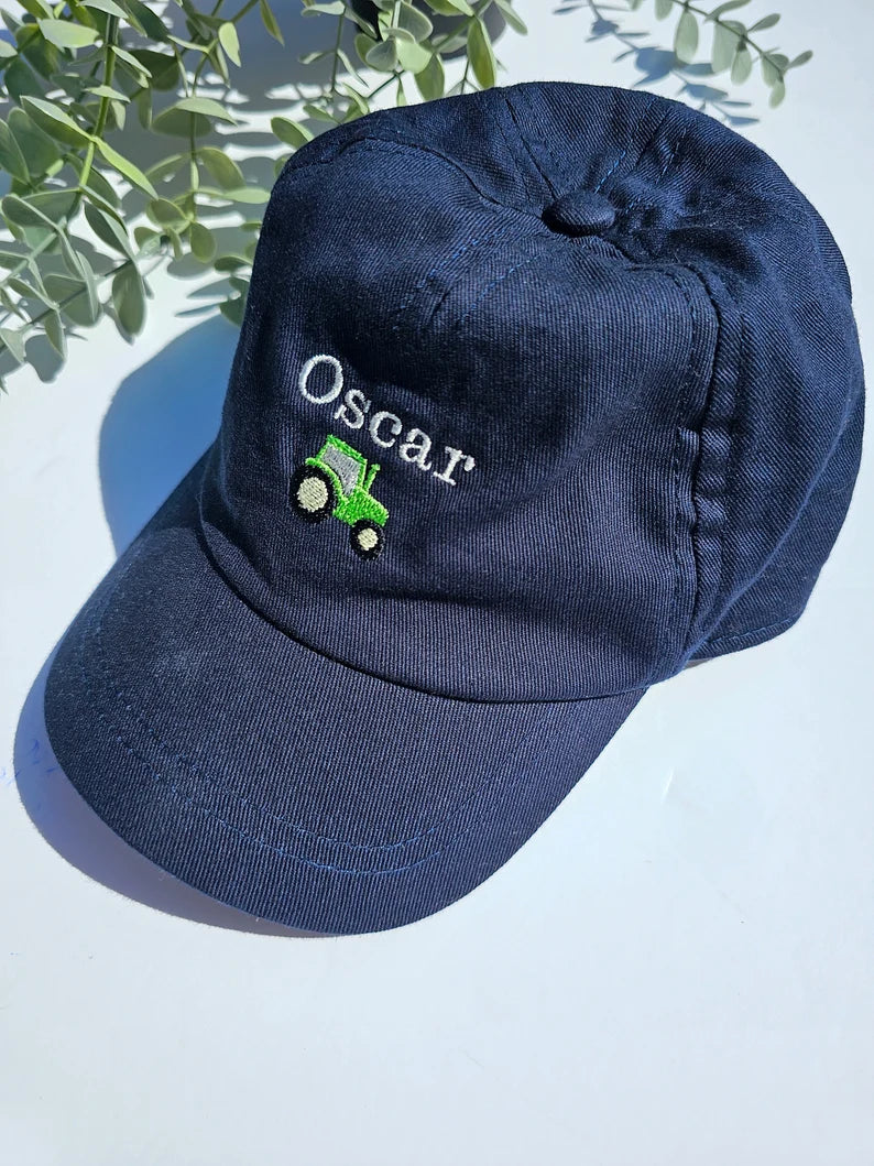 A navy blue toddler cap with the name 'Oscar' embroidered on the front, featuring a small green tractor below the name.