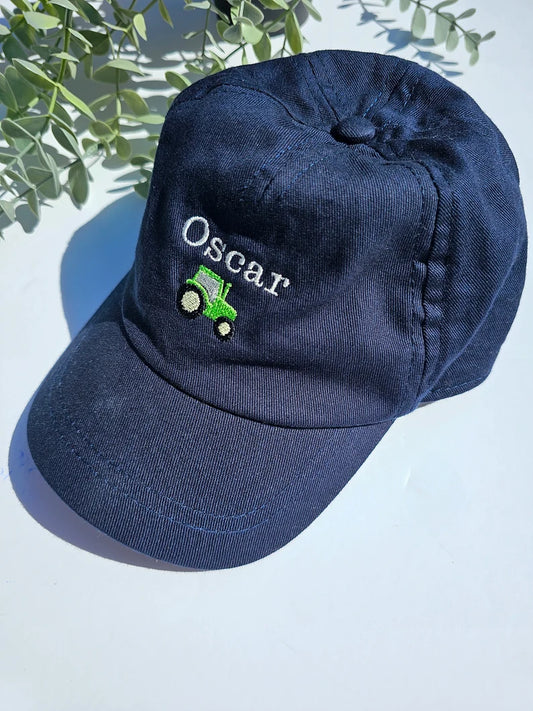 A navy blue toddler cap with the name 'Oscar' embroidered on the front, featuring a small green tractor below the name.