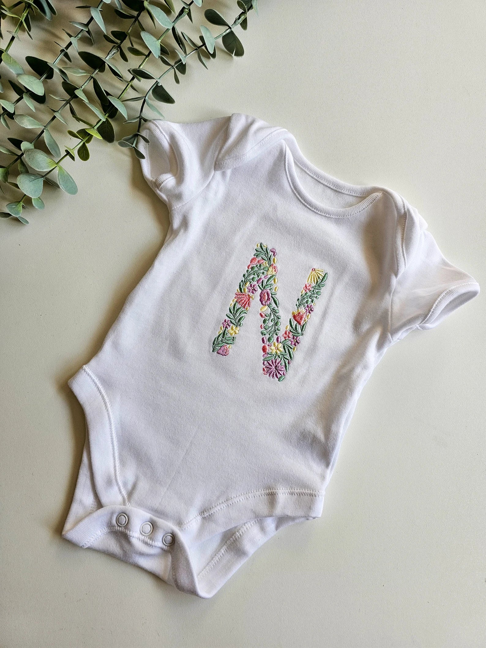 White baby onesie with floral letter design on a light background